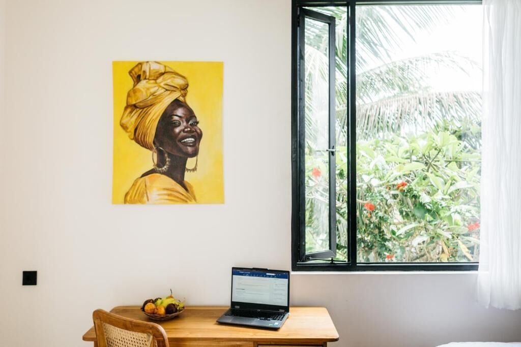 A room with a desk, a laptop, a chair, a fruit bowl, a window with a view of greenery, and a painting on the wall.