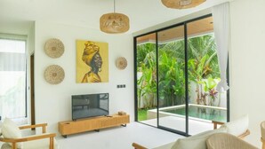 A modern living room with a TV, wooden furniture, and a large glass door leading to a pool. - Santara by Prime Villas (Ubud)