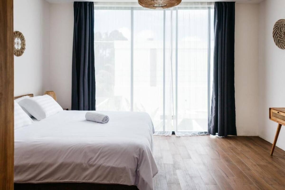 Un dormitorio con cama, piso de madera, ventana con cortinas y lámpara de pared.