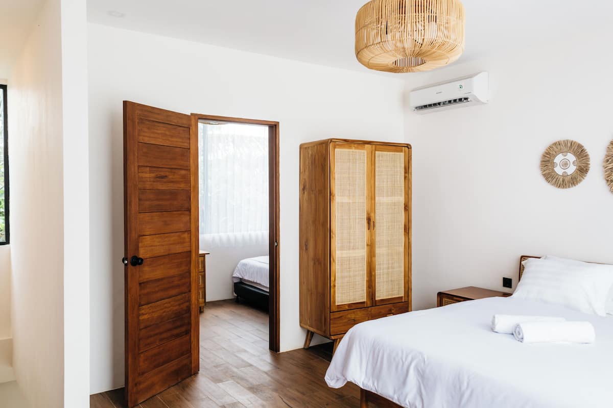 Un dormitorio con un armario de madera, una cama con ropa blanca, una lámpara colgante de mimbre y un equipo de aire acondicionado instalado en la pared.