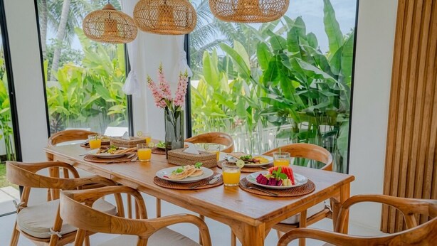 A wooden dining table with chairs, a vase with flowers, and a view of lush greenery through large windows. - Santara by Prime Villas (Ubud)