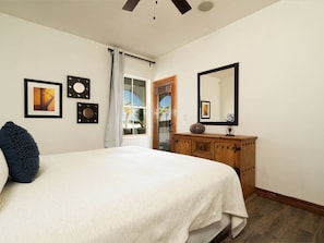A bedroom with a bed, a wooden dresser, a large mirror, and a window with a view of palm trees.