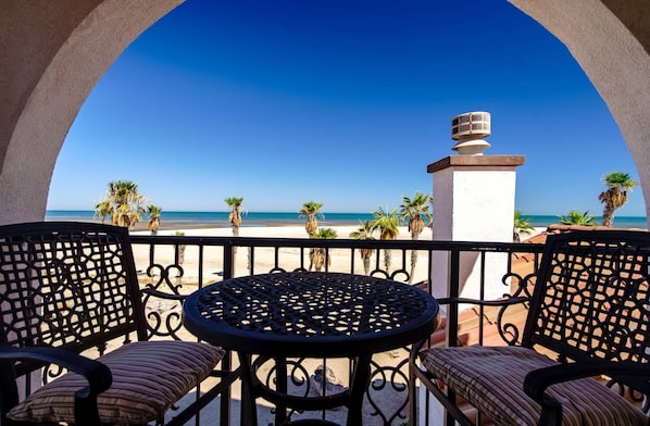 A balcony with a round table and chairs, overlooking a beach with palm trees and a clear blue sky.