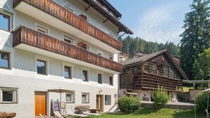 Un immeuble à plusieurs étages doté de balcons en bois, d’un espace extérieur couvert aménagé en coin salon et offrant une vue sur une colline boisée.