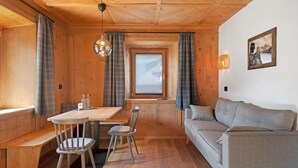 L’intérieur d’un chalet accueillant, avec une table à manger en bois, deux chaises, un banc, un canapé et une fenêtre avec des rideaux.