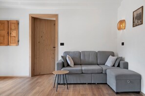 A modern living room with a grey sectional sofa, a small round table, and a wooden door. - Ferienwohnung 'Verena 2 – im Herzen der Dolomiten' (Urtijëi)