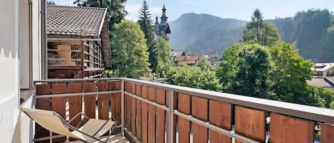 A balcony with a wooden railing, a folding chair, and a view of a mountain and village.