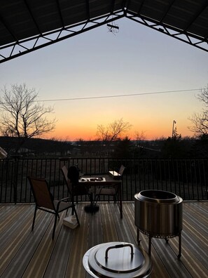 A wooden deck with a table and chairs, a metal grill, and a view of the sunset. - Unique Loft Rental with Access to Spacious Deck and Games Room for an Amazing Getaway by Tenkiller Ferry Lake in Oklahoma (Cookson)