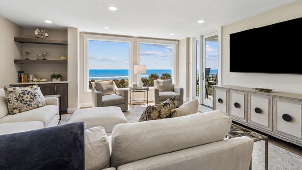 A living room with a large TV, comfortable seating, a bookshelf, and a view of the ocean. - 1368 Shipwatch - Hosted by Moore AI Rentals (Fernandina Beach)