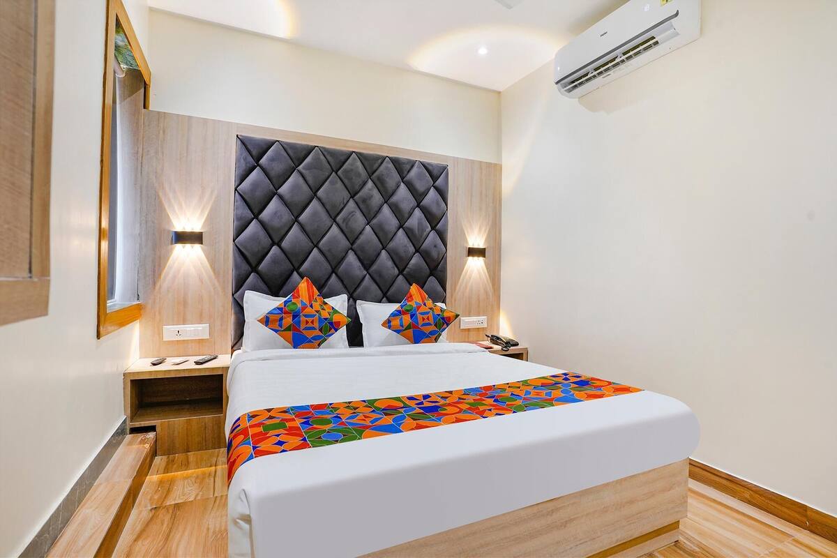 A hotel room with a tufted headboard, a bed with a colorful geometric-patterned bedspread, bedside tables with lamps, and an air conditioning unit.