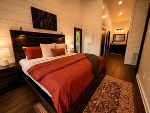 A bedroom with a large bed, a nightstand, a headboard, a wall-mounted artwork, a door leading to another room, and a patterned rug. - Cozy Cabin with Hot Tub & Fire Pit Easy Access to Nearby Lakes and Trails in Broken Bow, Oklahoma (Broken Bow)