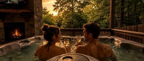 A hot tub with a stone fireplace, surrounded by a wooden deck and trees.