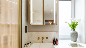 Ein Badezimmer mit einem weißen Waschbecken, goldenen Armaturen, einem Spiegel mit goldener Rahmung und einer Vase mit weißen Blumen auf der Ablagefläche.