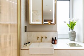 Ein Badezimmer mit einem weißen Waschbecken, goldenen Armaturen, einem Spiegel mit goldener Rahmung und einer Vase mit weißen Blumen auf der Ablagefläche.