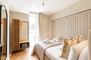 Ein Schlafzimmer mit einem Bett, einem Holzschrank, einer Bank, einem Spiegel und einem Fenster mit Vorhängen.