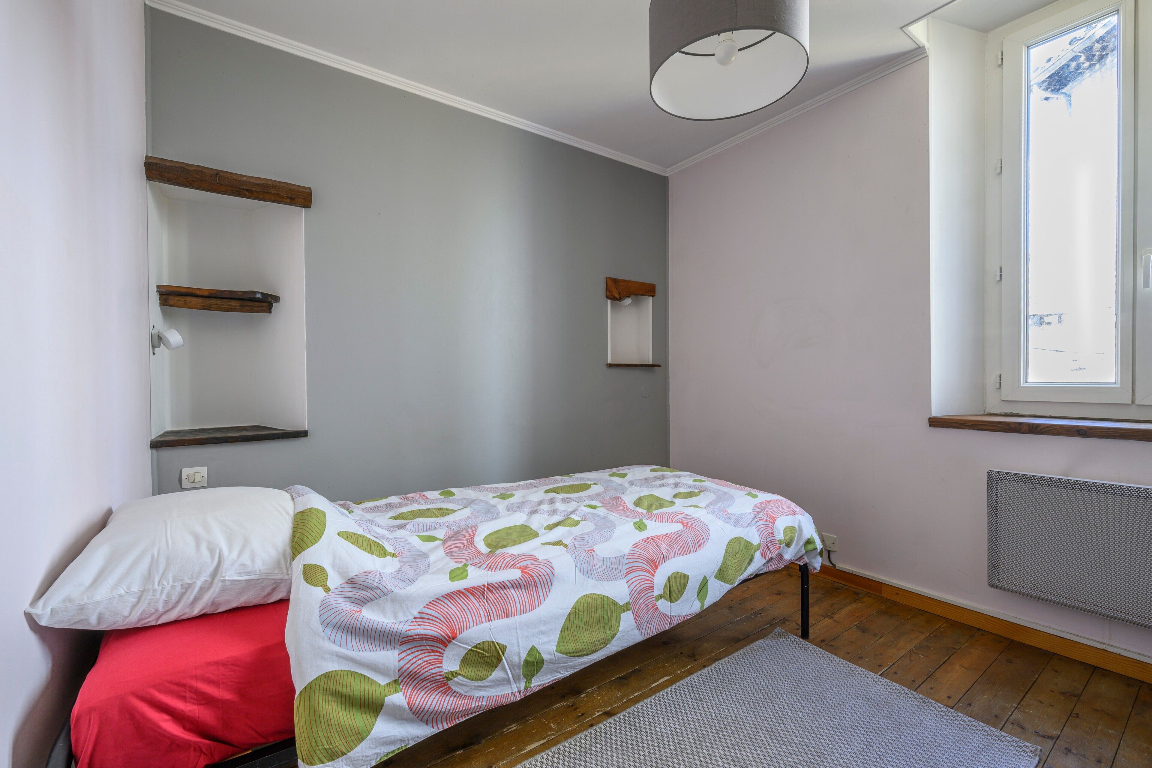 Une chambre à coucher avec un lit, un oreiller rouge, un oreiller blanc, une fenêtre avec des stores, un radiateur et un luminaire au plafond.