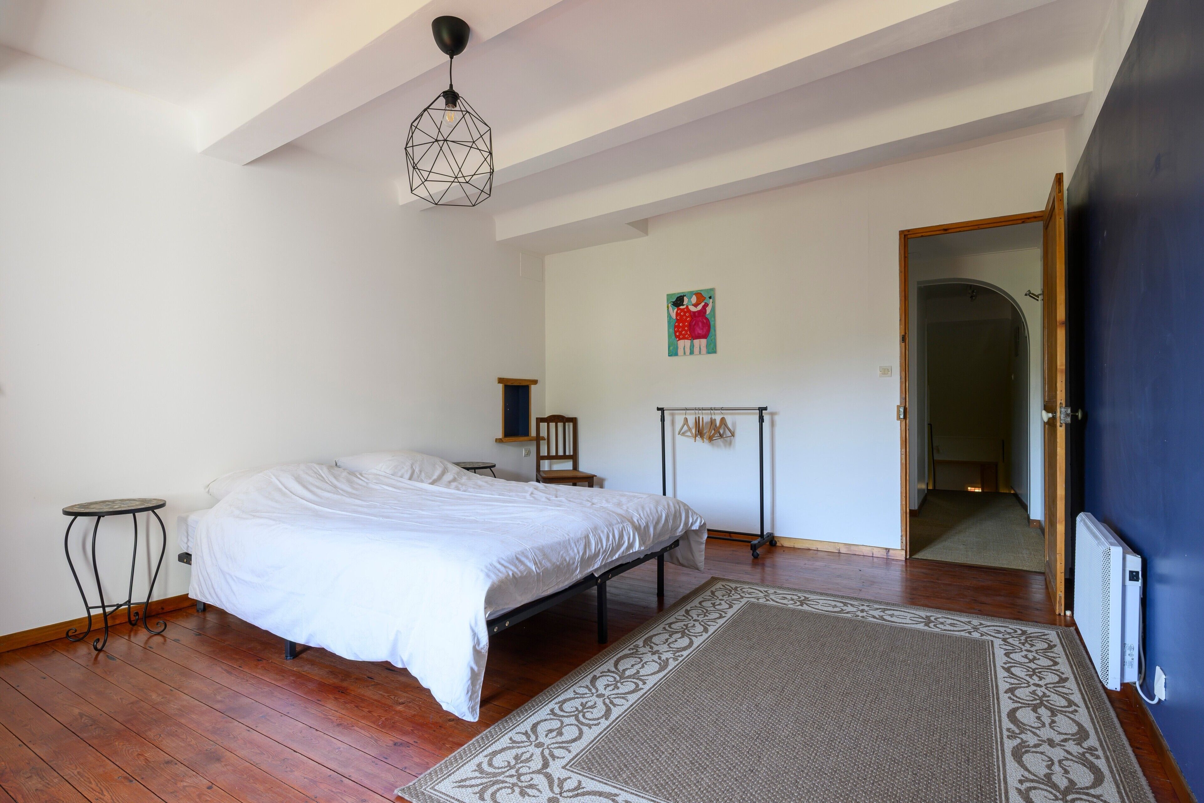 Een slaapkamer met een bed, een tafeltje, een radiator, een deur en een muur met een foto.