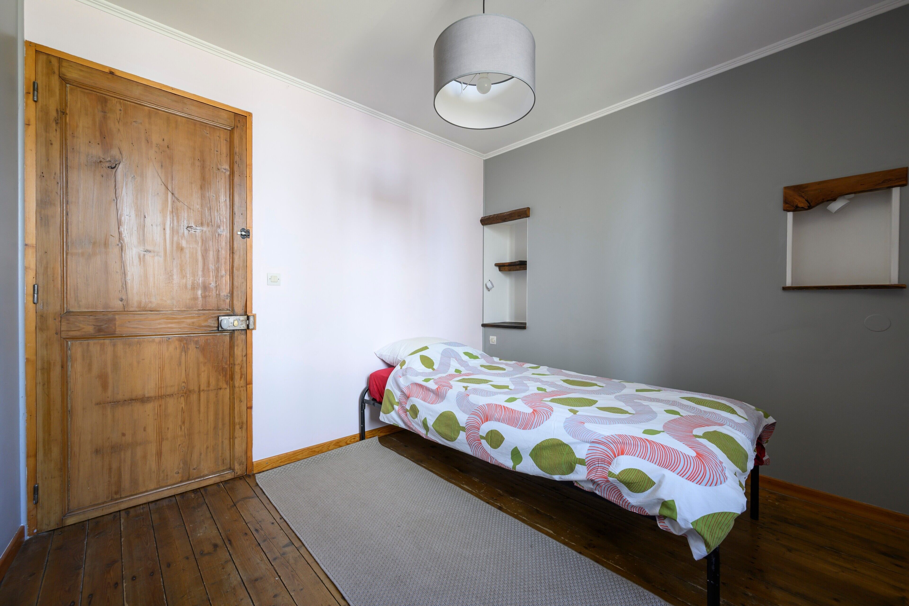 Een slaapkamer met een houten deur, een bed met een beddengoed met een patroon, een hanglamp en een klein wandrek.