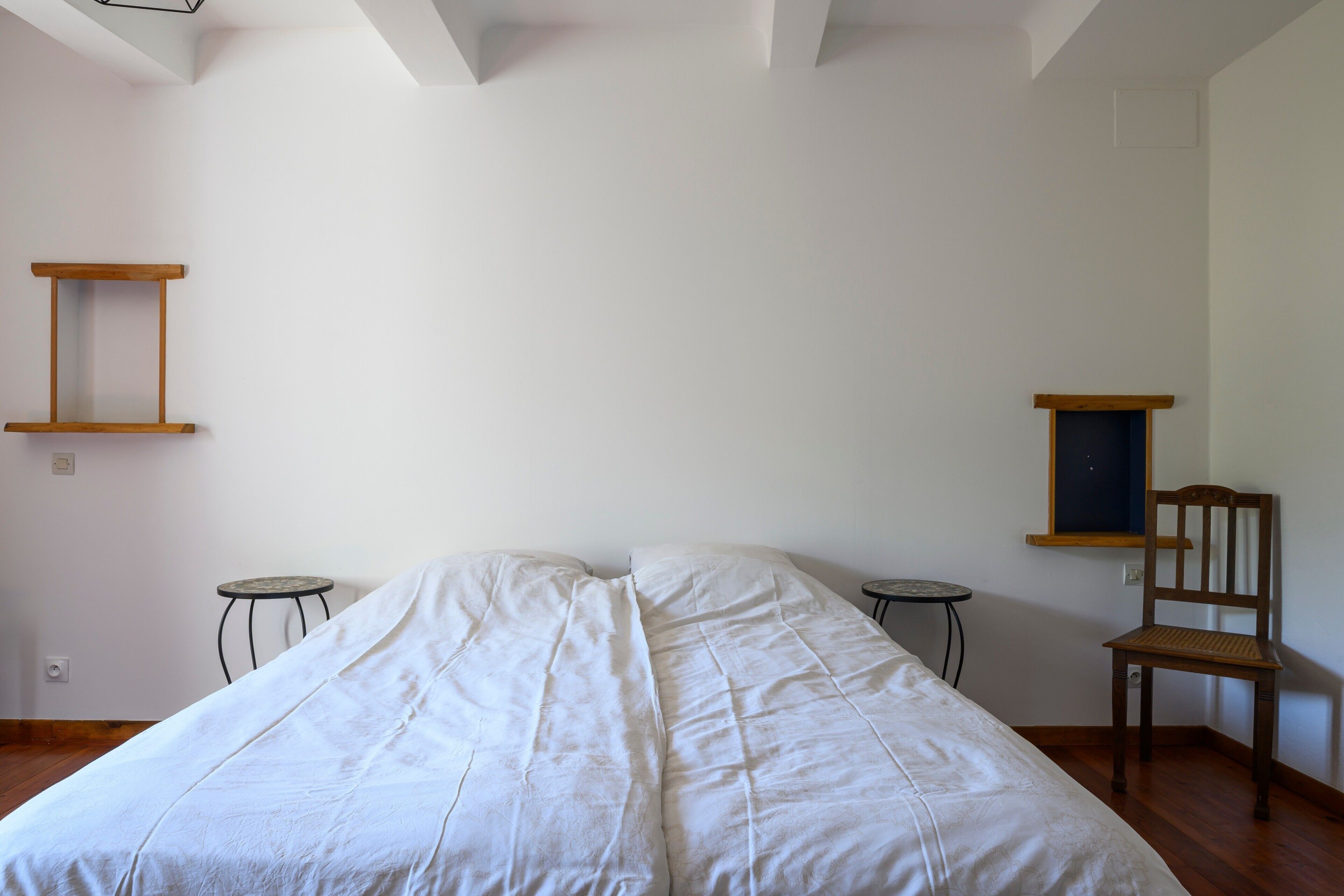 Un dormitorio con cama, dos mesitas pequeñas, una silla y un estante de madera en la pared.