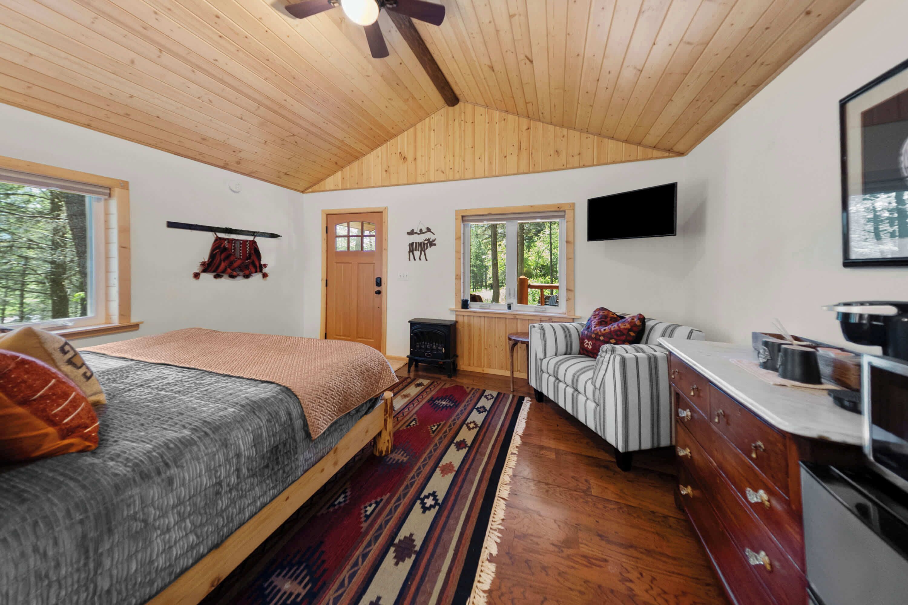 A bedroom with a bed, a sofa, a television, a wooden ceiling, and a window.
