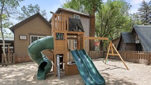 Ein Spielplatz mit Rutsche und Schaukeln, umgeben von einem Holzzaun und Bäumen.