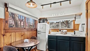 Eine Küche mit einem weißen Kühlschrank, einer Mikrowelle, einem Spülbecken und Holzschränken. Ein runder Tisch mit Stühlen und ein Fenster mit Blick auf Bäume.