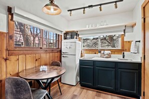 Une cuisine avec un réfrigérateur blanc, un four à micro-ondes, un évier et des armoires en bois. On y trouve une table ronde avec des chaises et une fenêtre donnant sur des arbres.