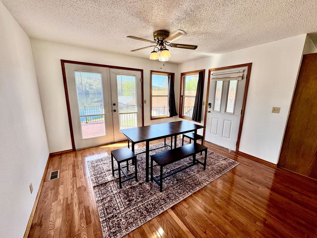 Una habitación con piso de madera, ventilador de techo, mesa de comedor con bancos y un tapete estampado.