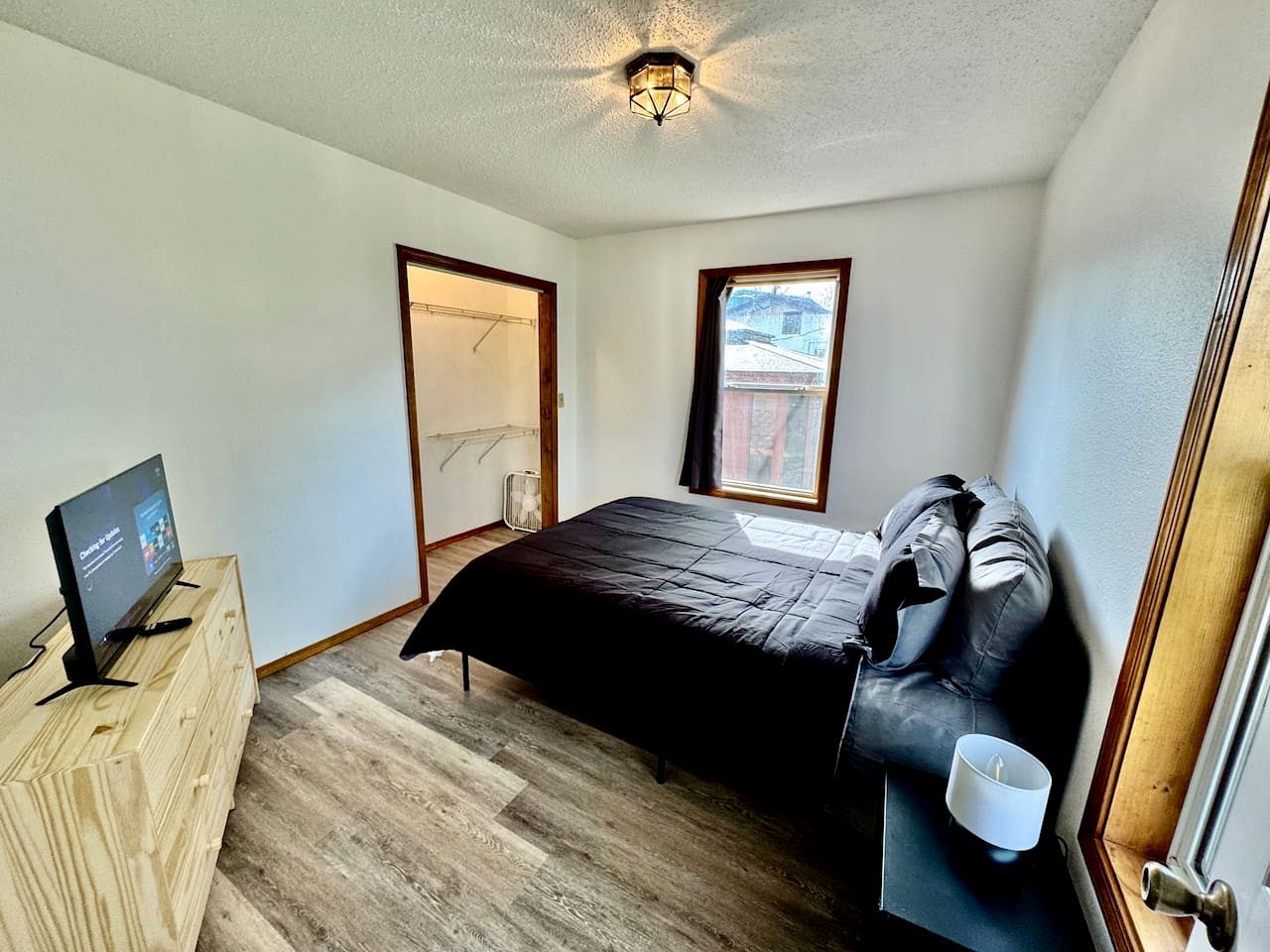 A bedroom with a bed, a TV on a wooden dresser, a window, and a door.
