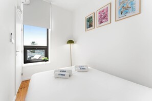 Ein schlichter Schlafzimmer mit einem großen Fenster, einem Bett mit zwei Kissen und zwei gerahmten Kunstwerken an der Wand.