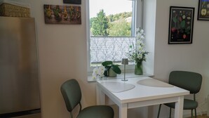 A small kitchen with a white table, two green chairs, a refrigerator, and a window with a view of trees.