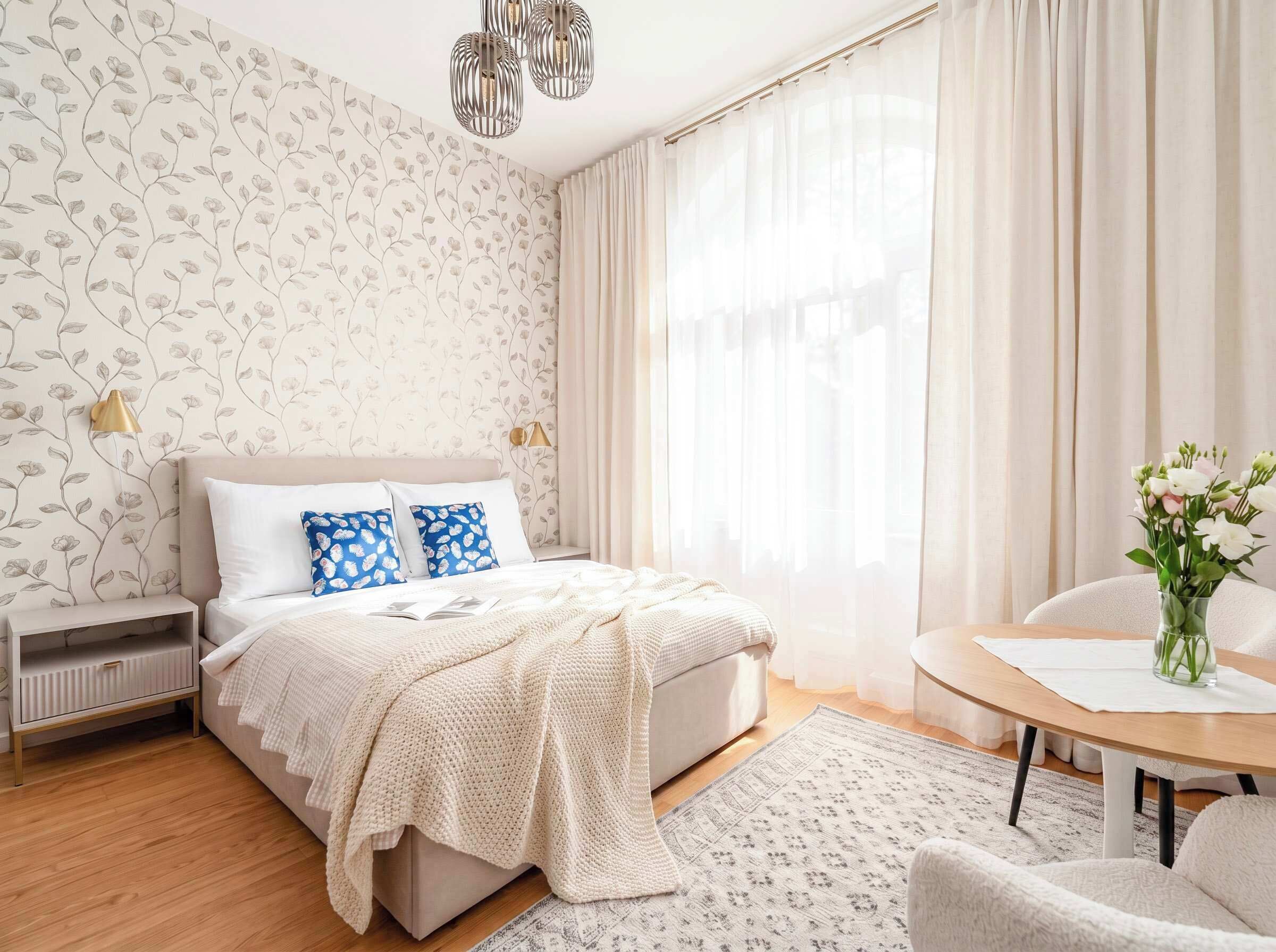 A bedroom with a bed, bedside lamps, a bedside table, a chair, a round table, and a vase with flowers.