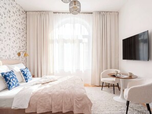 A hotel room with a bed, a desk, a chair, a TV, and a window with curtains. - TurnKey | Vinohrady Modern Studio with Balcony III (Prague 3)