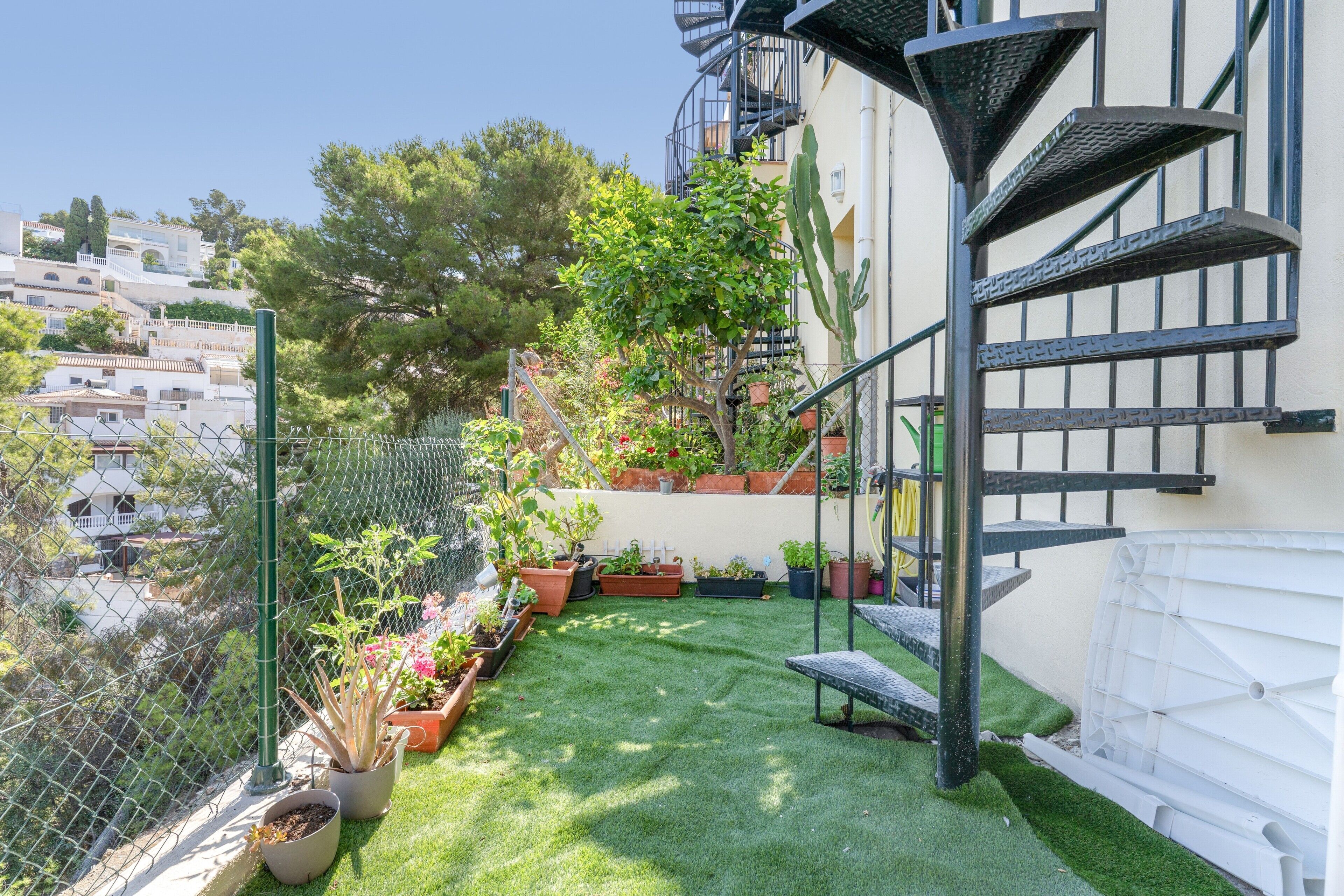 Un jardín en la azotea con plantas en macetas, una escalera espiral de metal y vistas de edificios circundantes.
