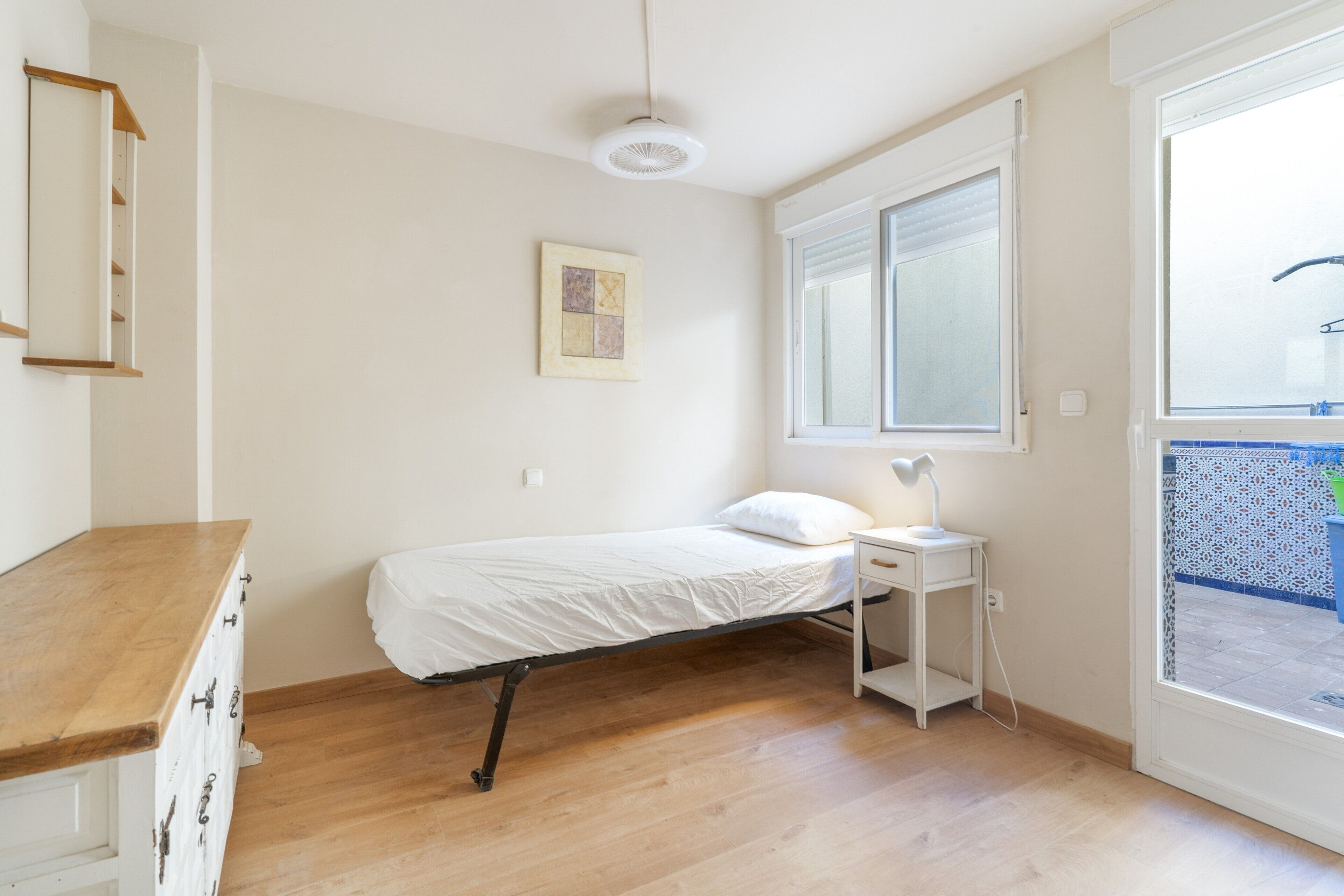 A single bed with a white mattress and pillow, a bedside table with a lamp, a window with blinds, and a wooden floor.
