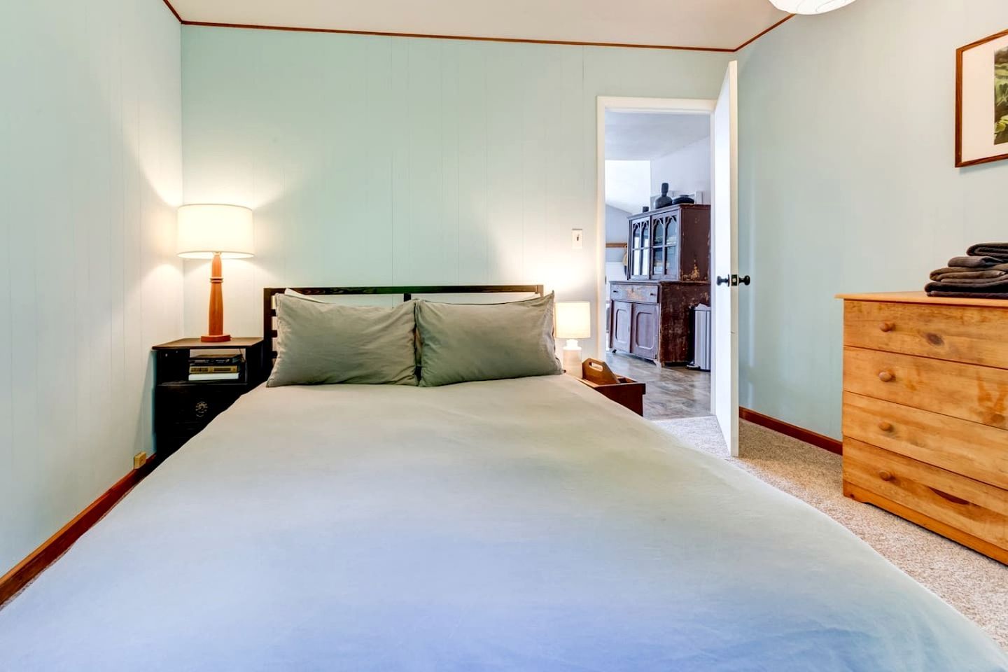 A bedroom with a bed, bedside table, lamp, dresser, and a door leading to another room.