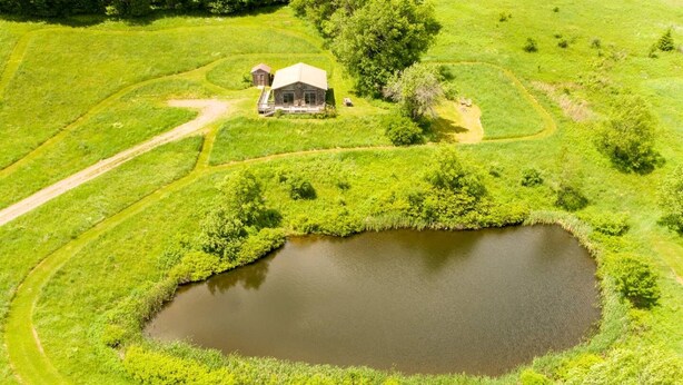 Een landelijk landschap met een vijver, een houten hutje en omliggende velden.
