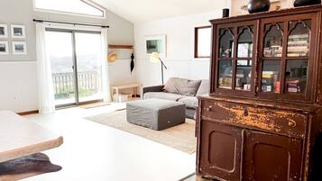 A living room with a sofa, ottoman, a wooden cabinet, a table, and a large window with a view.