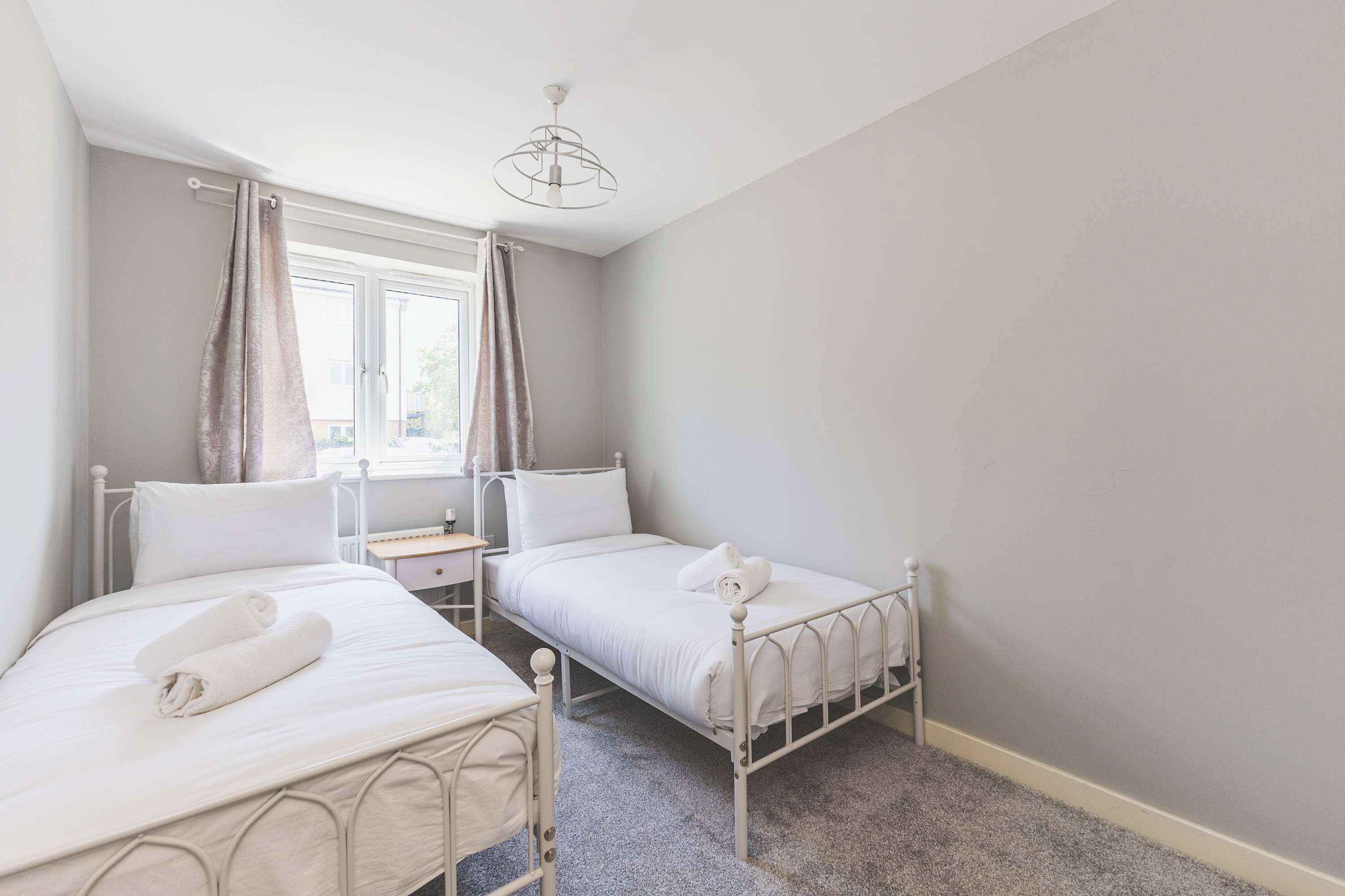 A bedroom with two single beds, a window with curtains, a bedside table, and a hanging light fixture.