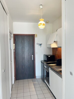 Ein schmaler Flur, der zu einer Küche mit Mikrowelle, Backofen und einer Pendelleuchte führt.
