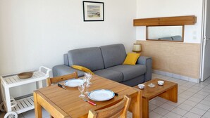 Um quarto com um sofá cinza, mesa de jantar de madeira e cadeiras, um carrinho branco pequeno com uma tigela, e um quadro pendurado na parede.