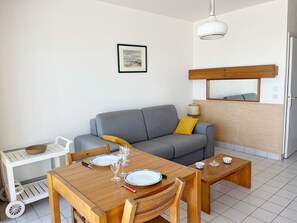 Ein Zimmer mit grauem Sofa, einem Holzesstisch und Stühlen, einem kleinen weißen Tablett mit einer Schüssel und einem gerahmten Bild an der Wand.