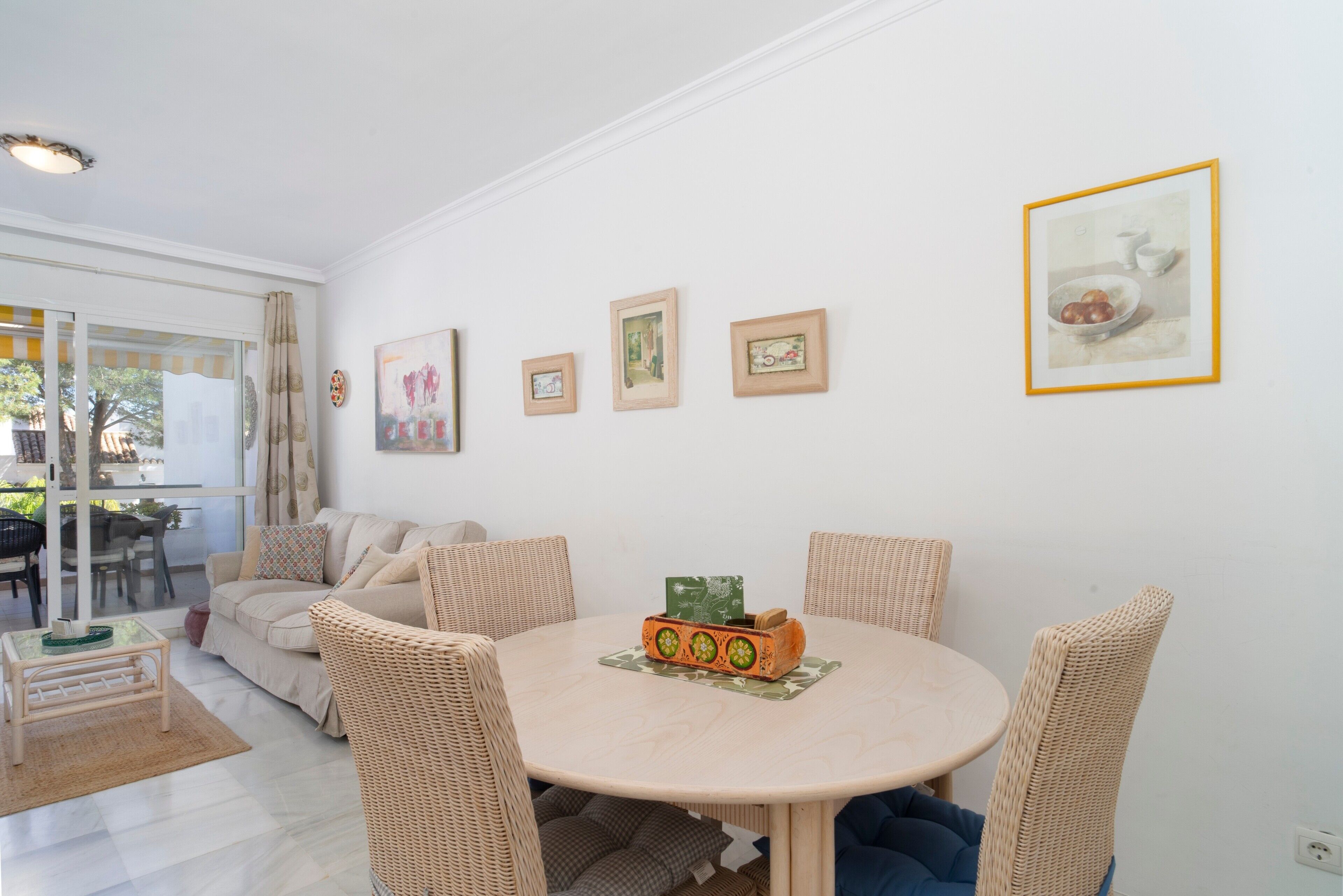 Ein Wohnzimmer mit rundem Tisch, Korbstühlen, einer Couch und gerahmten Bildern an der Wand.