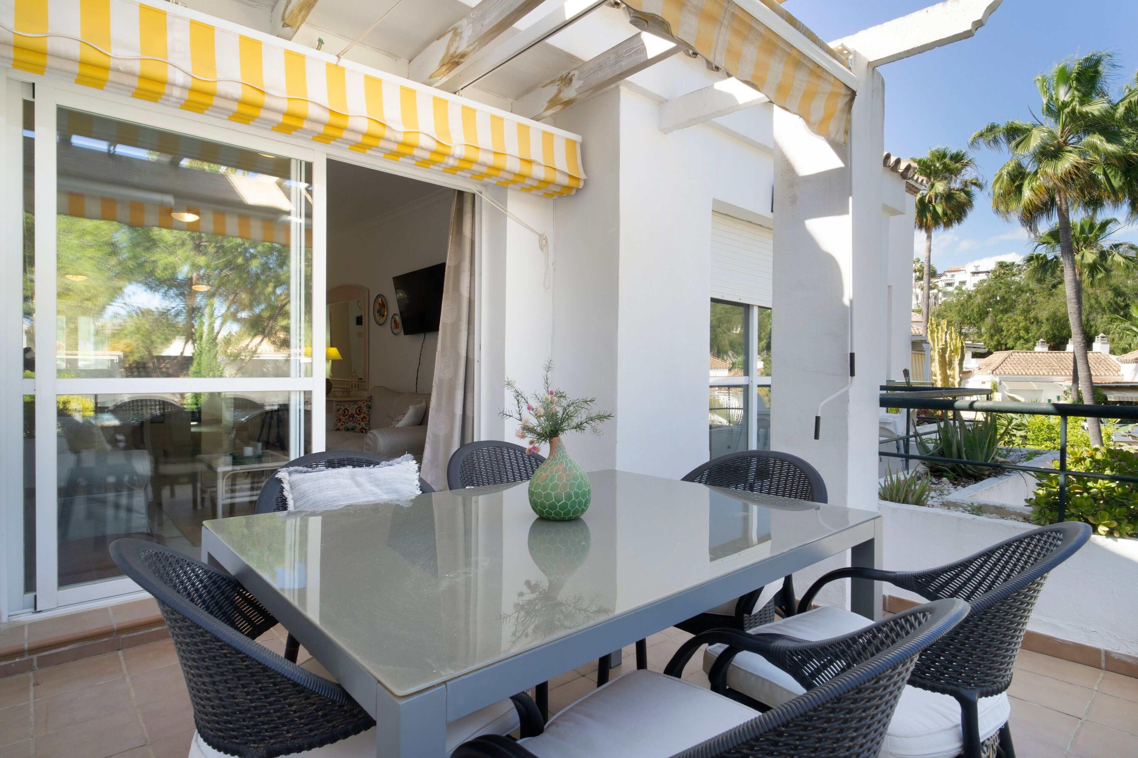 Un patio con una mesa de vidrio y sillas de mimbre, un toldo amarillo y blanco, y vistas a palmeras y un edificio al fondo.