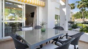 Un patio con una mesa de vidrio y sillas de mimbre, un toldo amarillo y blanco, y vistas a palmeras y un edificio al fondo.