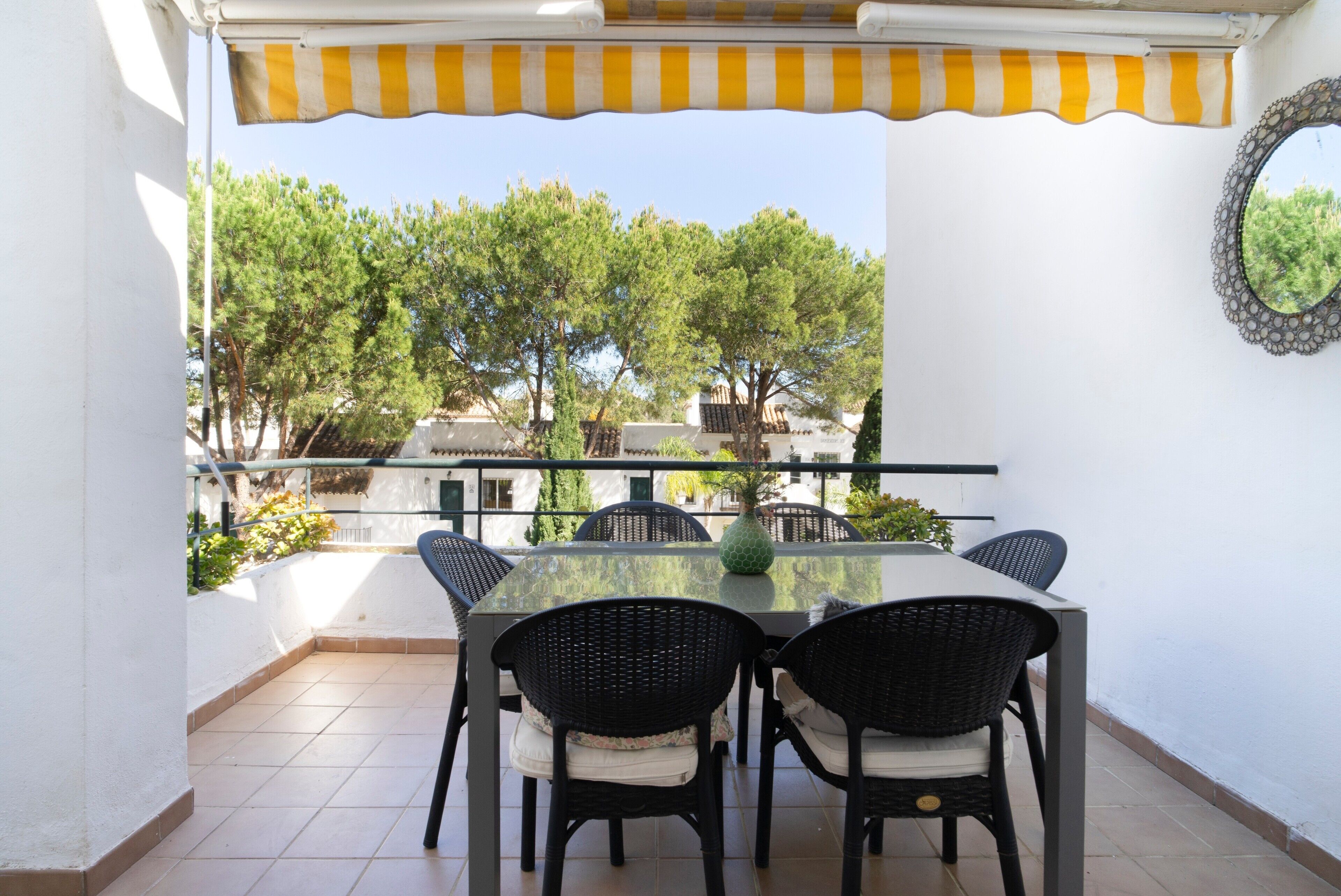 A covered outdoor dining area with a table and chairs, a yellow and white striped awning, and a view of trees and buildings.