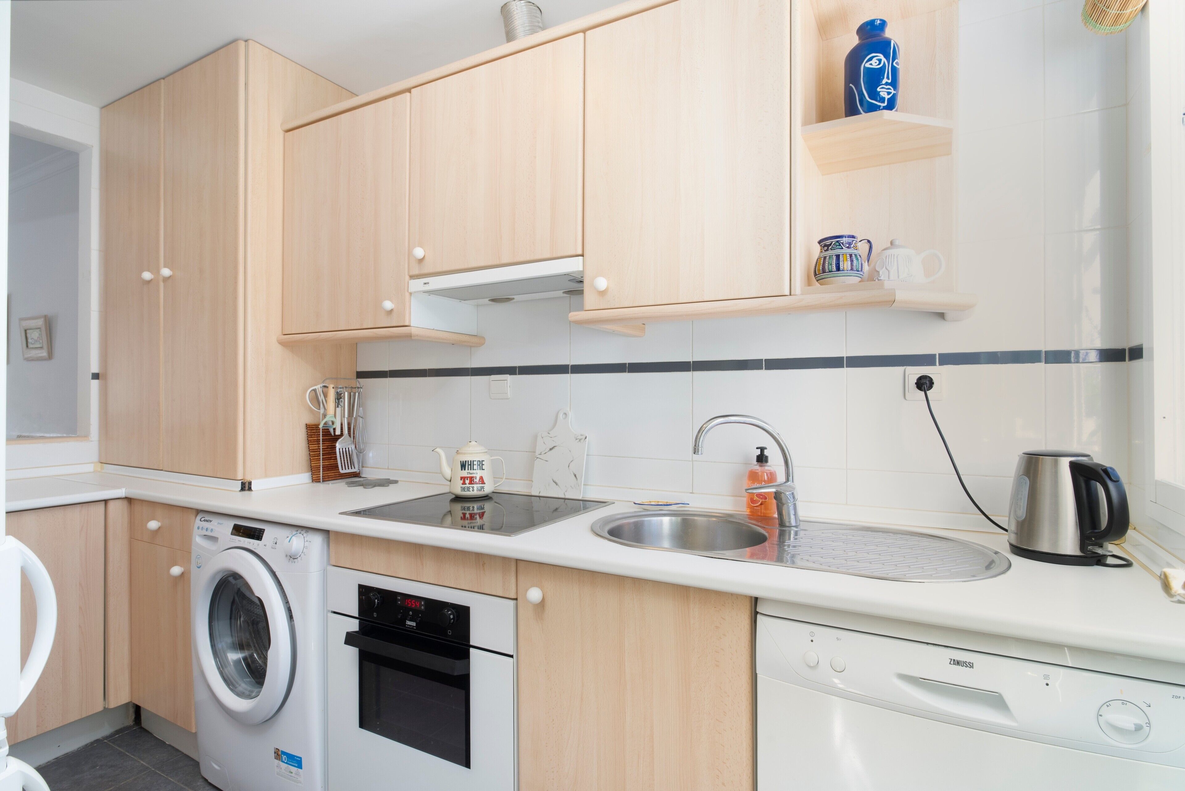 A modern kitchen with wooden cabinets, a washing machine, an oven, a dishwasher, a sink, and a kettle.