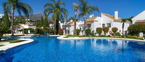 A swimming pool surrounded by white buildings with red-tiled roofs, palm trees, and a clear blue sky.