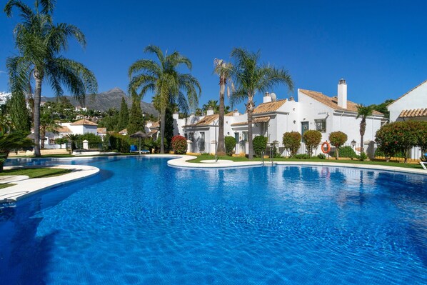 A swimming pool surrounded by white buildings with red-tiled roofs, palm trees, and a clear blue sky. - Vibrant Golf Valley Apartment with Terrace and Three Pools (Marbella)