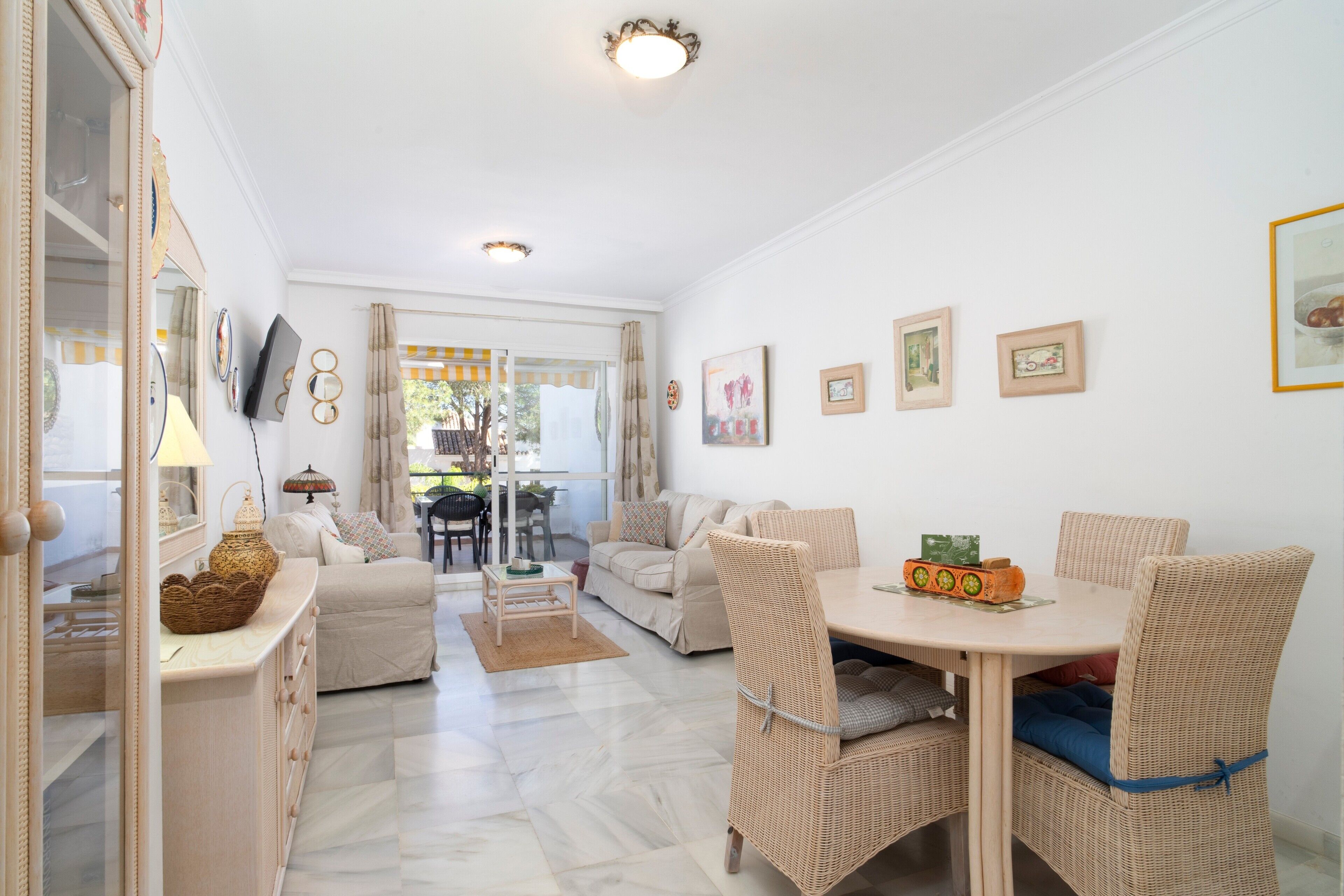 A well-lit, spacious living area with a dining table, wicker chairs, a sofa, and a TV.